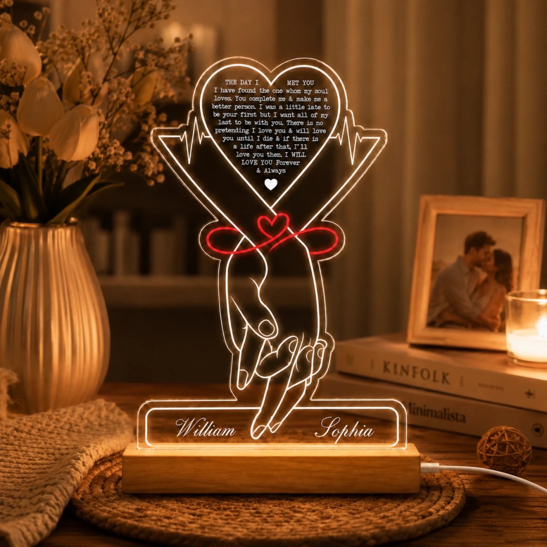 A glowing acrylic LED lamp depicting two hands tied with a red ribbon and a heart, with names "William" and "Sophia" on the base. It sits on a table with a vase of white flowers, a framed photo, and a lit candle.