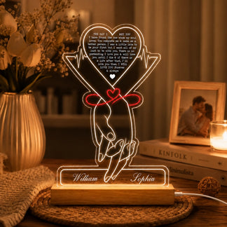 A glowing acrylic LED lamp depicting two hands tied with a red ribbon and a heart, with names "William" and "Sophia" on the base. It sits on a table with a vase of white flowers, a framed photo, and a lit candle.