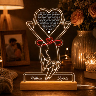 A glowing acrylic LED lamp featuring holding hands tied with a red ribbon and "William" and "Sophia" on its wooden base. It's on a nightstand with pink roses, a framed picture, and a candle.