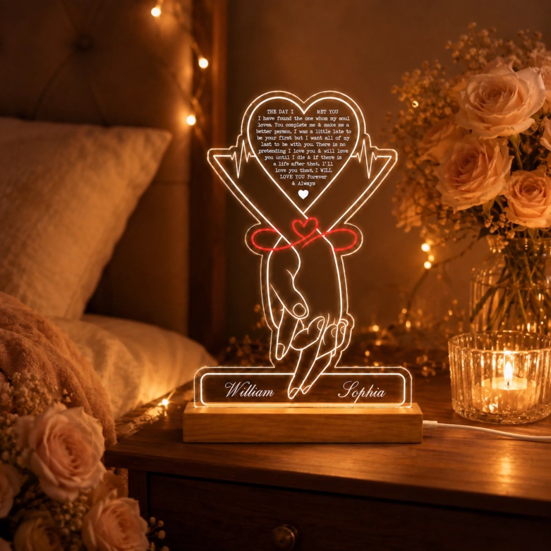 A glowing acrylic LED lamp showing holding hands with a red ribbon and "William" and "Sophia" on its base. It's on a bedside table surrounded by string lights, roses, and a lit candle.