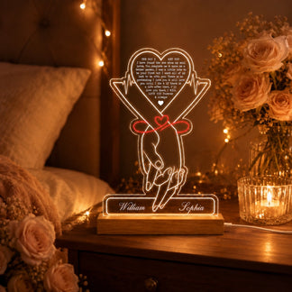 A glowing acrylic LED lamp showing holding hands with a red ribbon and "William" and "Sophia" on its base. It's on a bedside table surrounded by string lights, roses, and a lit candle.