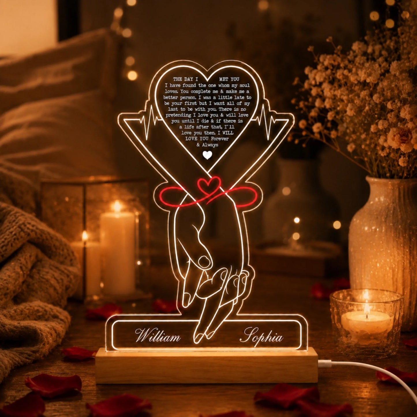 A glowing acrylic LED lamp depicting two holding hands with a red ribbon and "William" and "Sophia" on its wooden base. It's surrounded by white candles, flowers, and scattered red rose petals.