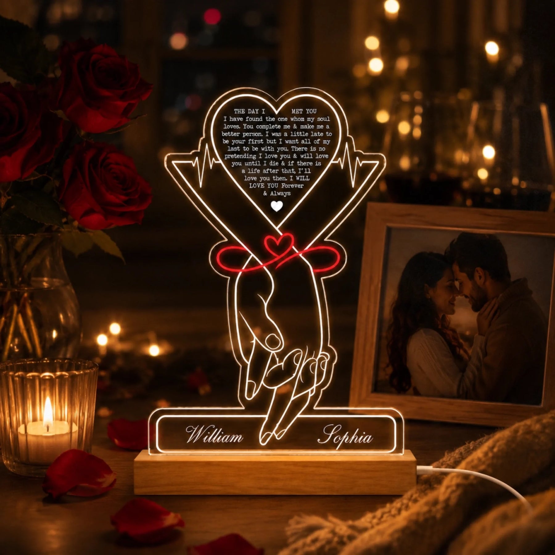 A glowing acrylic LED lamp with holding hands tied by a red ribbon and "William" and "Sophia" on its base. It's in a romantic setting with red roses, a framed photo, and candle light.