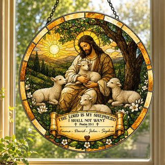 A circular stained-glass style suncatcher hanging in a window, depicting a robed shepherd figure embracing a lamb, surrounded by three other lambs, under a sun. A banner below reads "Emma - David - John - Sophia".