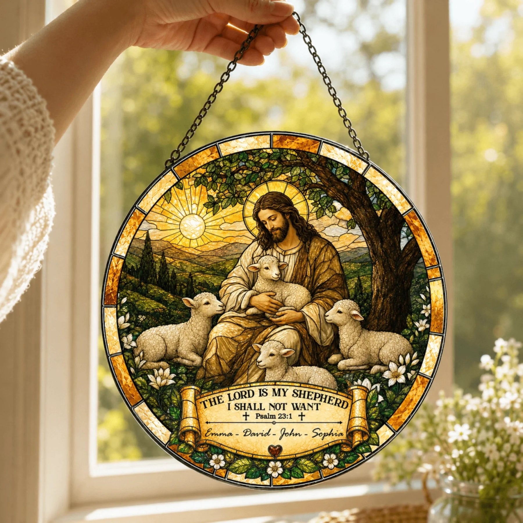 A hand holds a circular stained-glass style suncatcher featuring a robed shepherd figure with lambs, in front of a bright window. A banner reads "Emma - David - John - Sophia".
