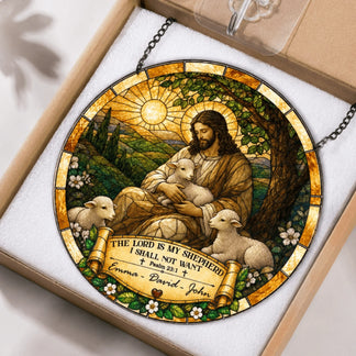 A circular stained-glass style suncatcher, depicting a robed shepherd figure with lambs, rests in an open brown cardboard box with foam. A banner reads "Emma - David - John".
