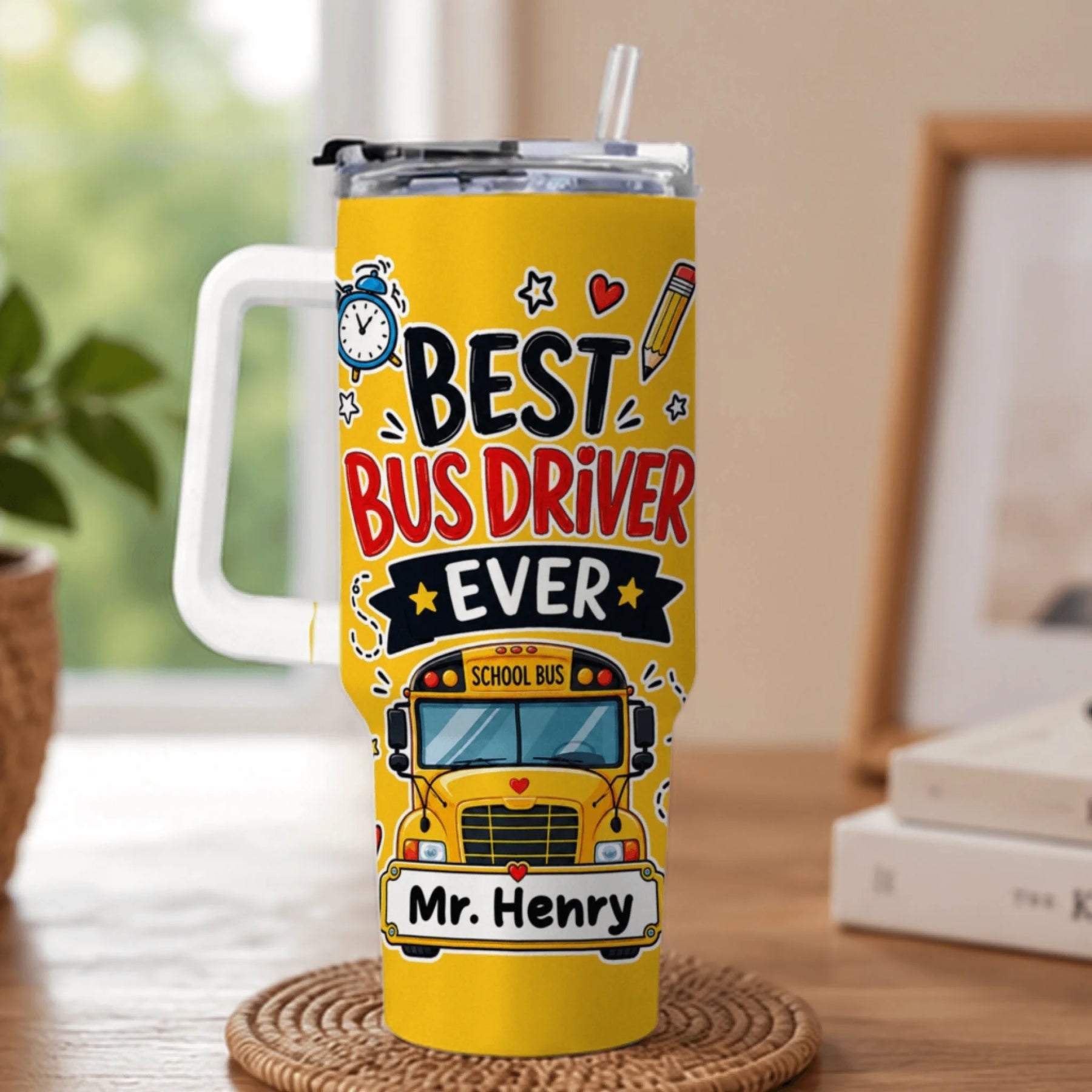 A yellow tumbler with a white handle and straw, printed with "BEST BUS DRIVER EVER", a school bus graphic, and "Mr. Henry" on a coaster.