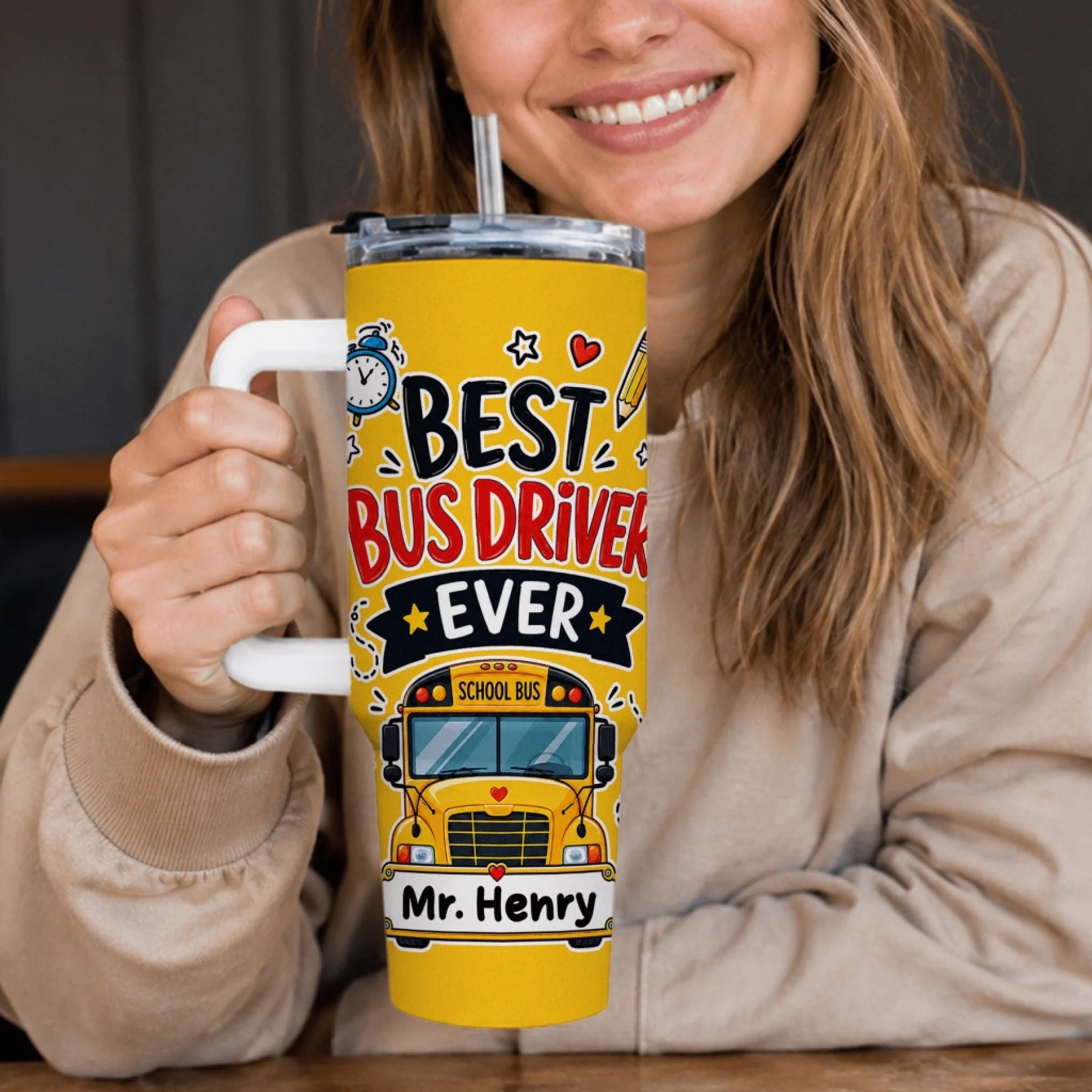 A smiling person in a beige top holds a yellow tumbler with a white handle. It says "BEST BUS DRIVER EVER" with a school bus and "Mr. Henry".