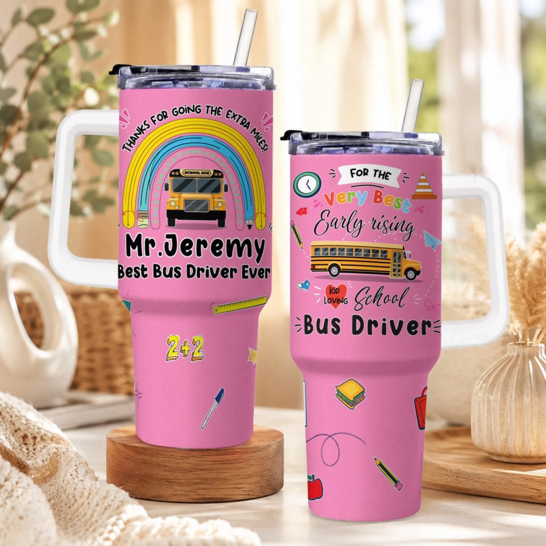 Two pink handled tumblers with straws, featuring school bus driver appreciation designs. The left tumbler has a rainbow and school bus.