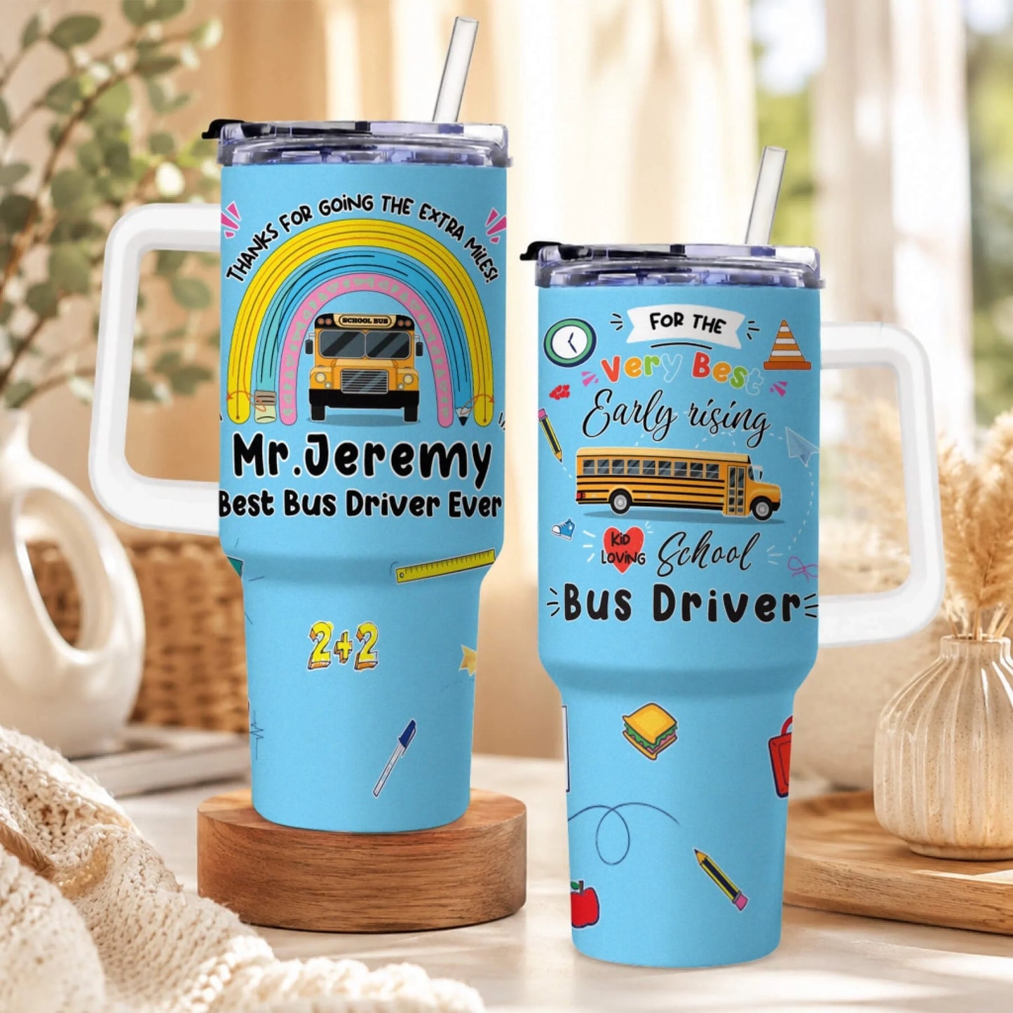 Two light blue handled tumblers with straws, featuring school bus driver appreciation designs. The left tumbler has a rainbow and school bus.