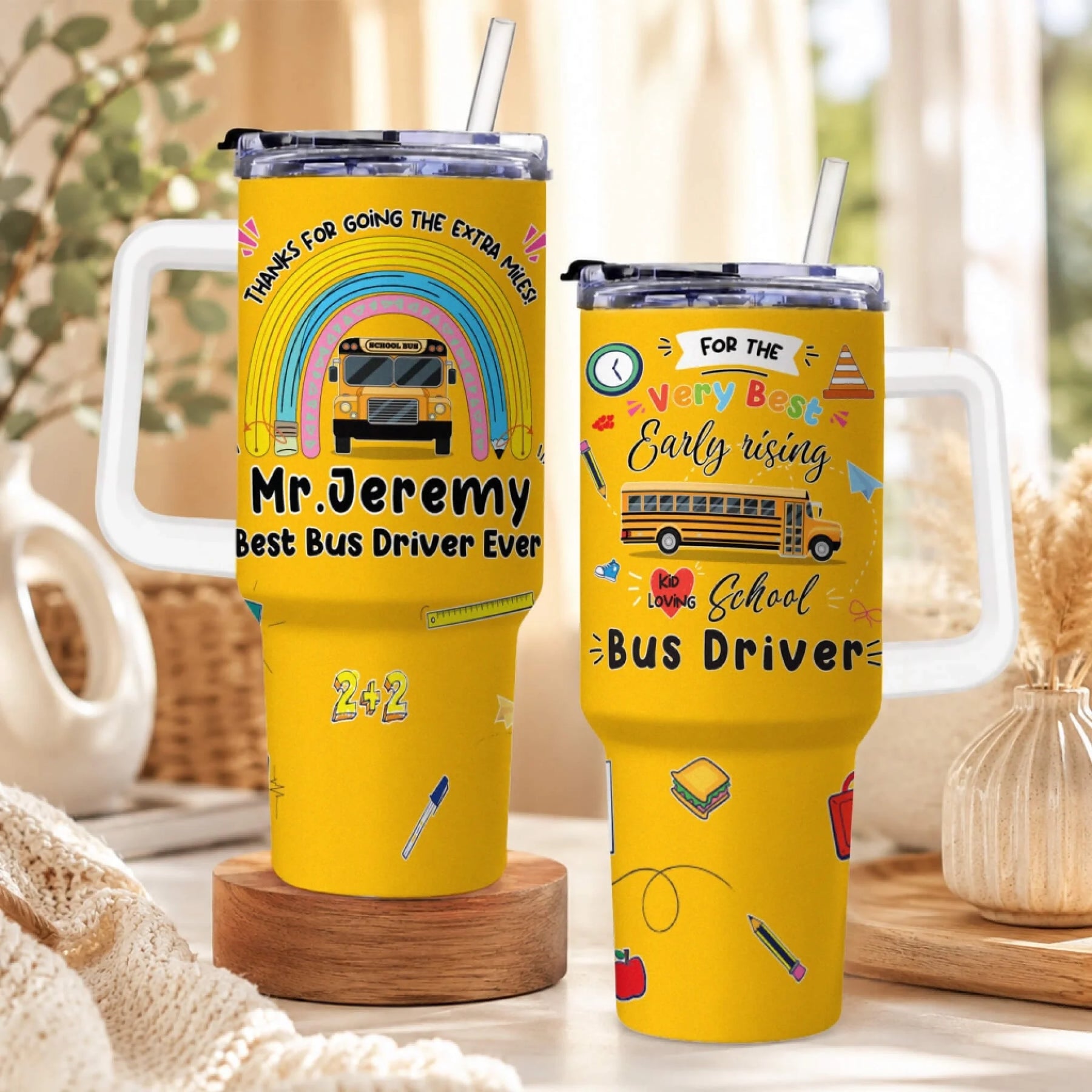 Two yellow handled tumblers with straws, featuring school bus driver appreciation designs. The left tumbler has a rainbow and school bus.