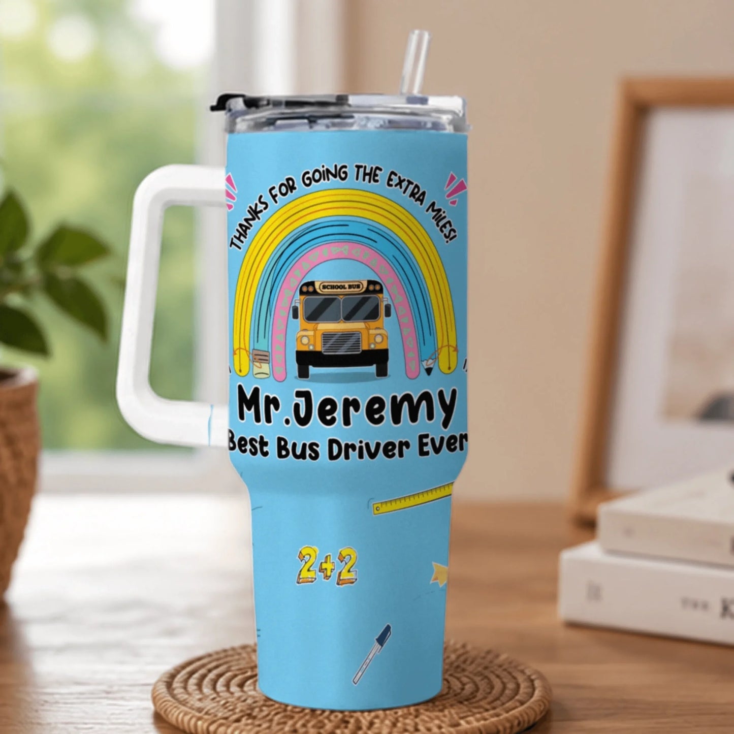 A light blue handled tumbler with a straw, adorned with a rainbow, school bus, and text appreciating a bus driver.