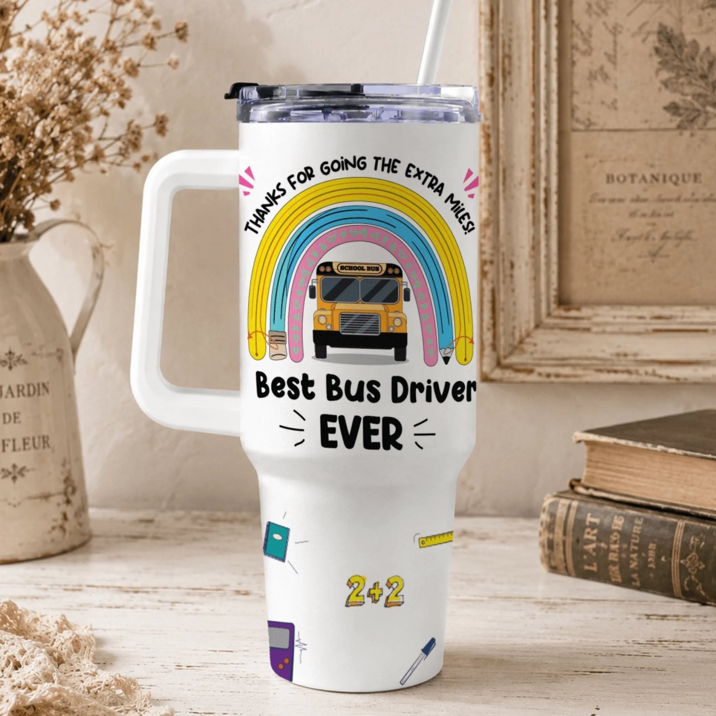 A white handled travel tumbler features a rainbow, school bus, and text "Best Bus Driver EVER" along with other school-themed graphics.