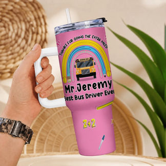 A hand holds a pink handled travel tumbler with a rainbow, school bus, and personalized text "Mr. Jeremy Best Bus Driver Ever".