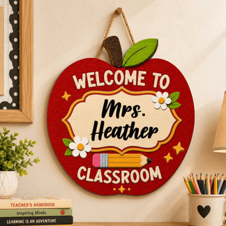 An apple-shaped wooden sign reading "WELCOME TO Mrs. Heather CLASSROOM" with a pencil and flowers, hanging on a wall.