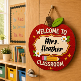 An apple-shaped wooden sign reading "WELCOME TO Mrs. Heather CLASSROOM" with a pencil and flowers, hanging on a door.