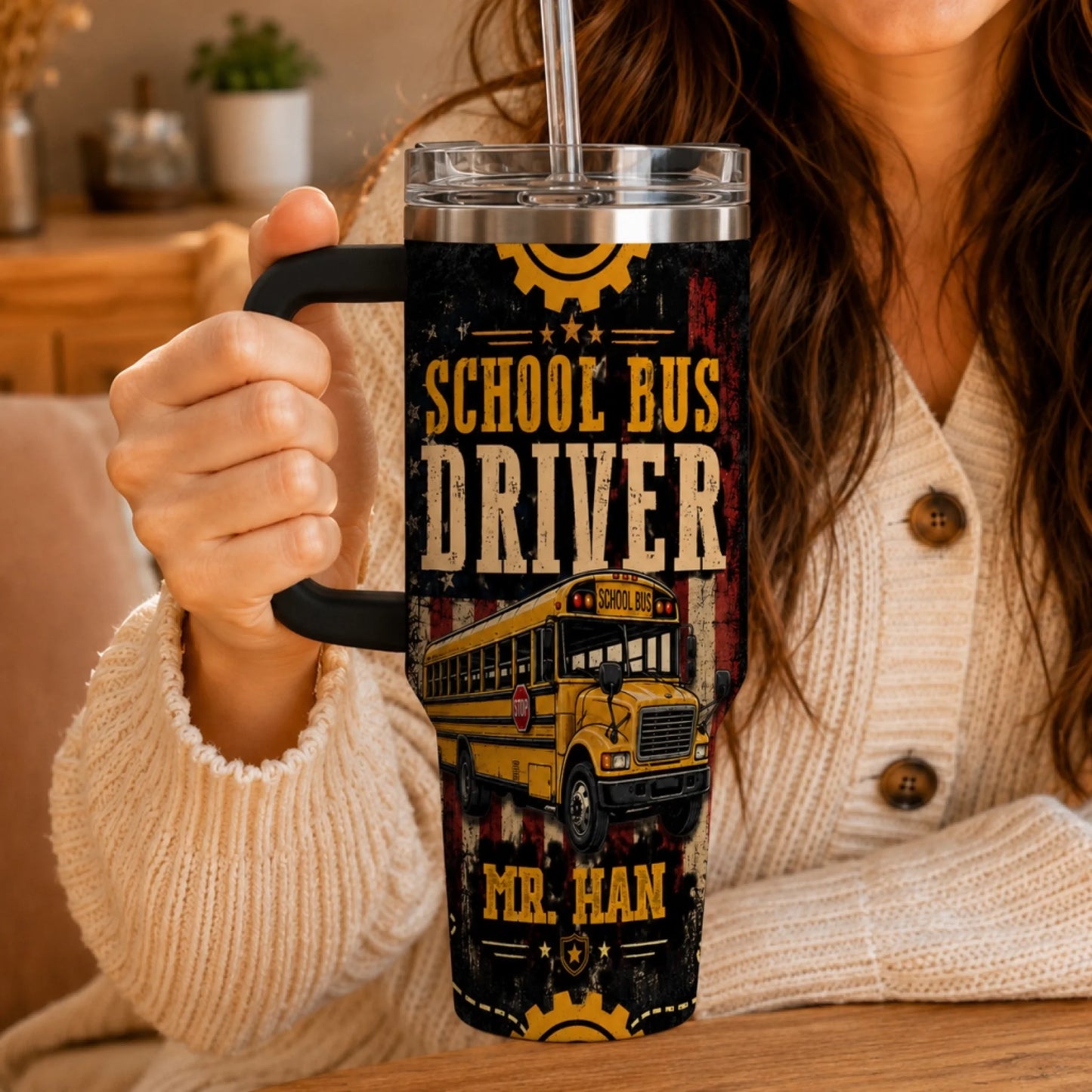 A person wearing a beige cardigan holds a handled tumbler with a straw, showing a yellow school bus design and 'School Bus Driver' text.