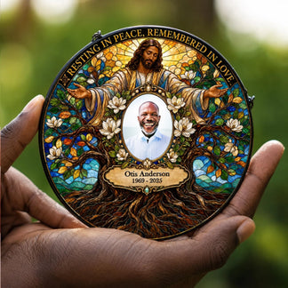 A personalized round memorial plaque held in two hands against a blurred green outdoor background. The plaque features a stained-glass-style design with an image of Jesus Christ, white magnolia flowers, and a central portrait of a smiling man labeled 'Otis Anderson 1969 - 2025'.