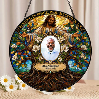 Personalized stained-glass-style hanging memorial plaque featuring Jesus with outstretched arms, white flowers, tree roots, and the text 'Resting in Peace, Remembered in Love,' with a circular portrait photo in the center and the name Otis Anderson 1969–2025 below.