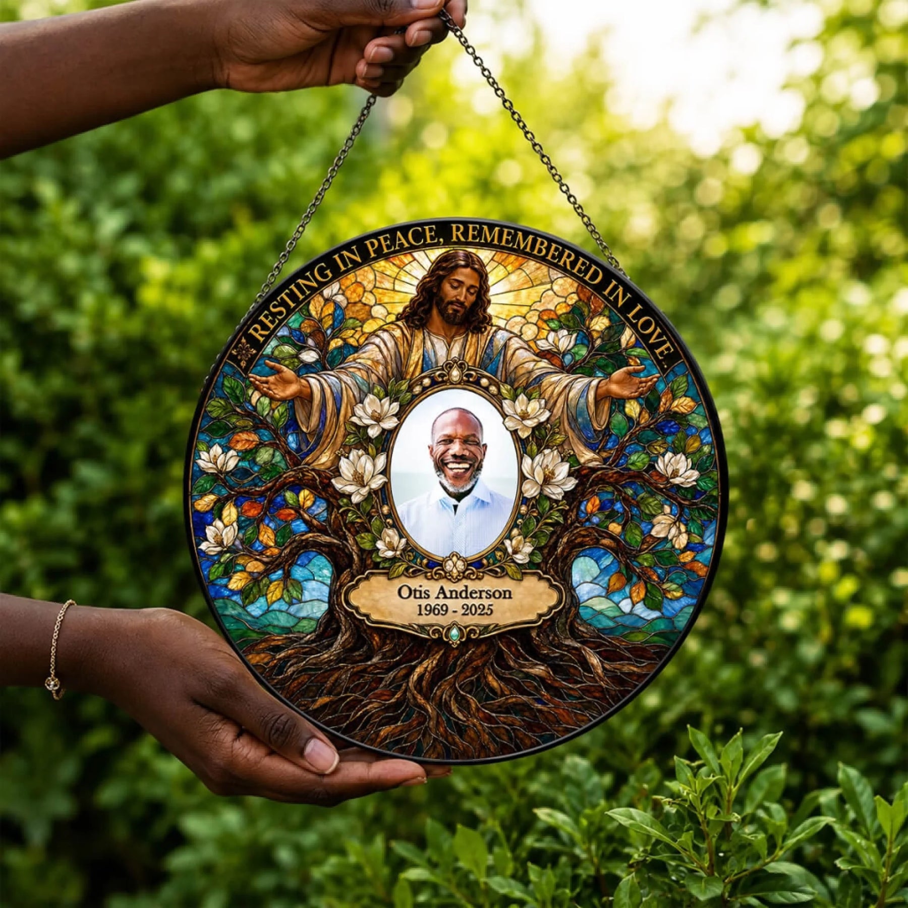A circular stained-glass-style memorial hanging plaque is held by two hands against a blurred green garden background. The plaque features an illustrated figure of Jesus surrounded by flowers and tree roots, with a central portrait of a smiling man and the text 'RESTING IN PEACE, REMEMBERED IN LOVE' and 'Otis Anderson 1969-2025'.