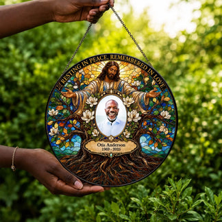 A circular stained-glass-style memorial hanging plaque is held by two hands against a blurred green garden background. The plaque features an illustrated figure of Jesus surrounded by flowers and tree roots, with a central portrait of a smiling man and the text 'RESTING IN PEACE, REMEMBERED IN LOVE' and 'Otis Anderson 1969-2025'.