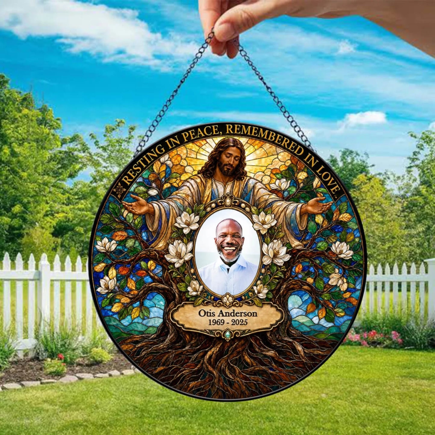 A circular hanging memorial plaque being held by a hand outdoors. The plaque features a stained-glass-style design with a central portrait of a smiling man, surrounded by a robed figure with outstretched arms, flowers, tree roots, and the text 'RESTING IN PEACE, REMEMBERED IN LOVE' and 'Otis Anderson 1969 - 2025.