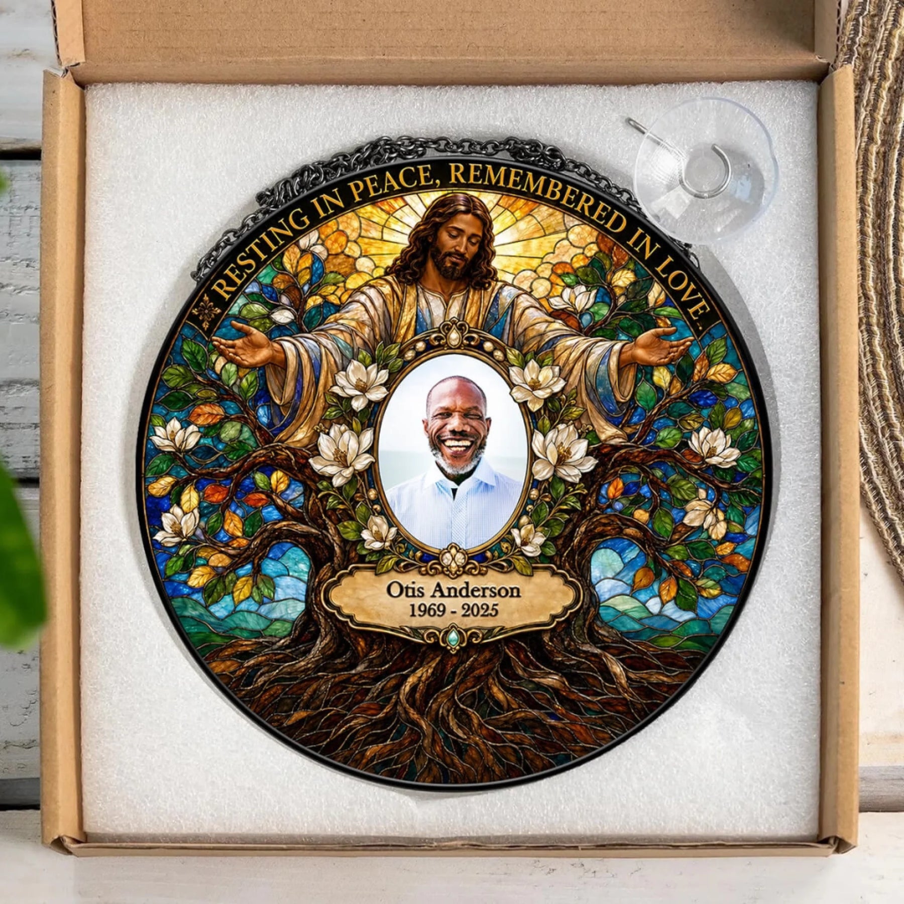 Personalized stained-glass style memorial plate displayed inside a cardboard gift box. The circular plate features an illustrated figure of Jesus with outstretched hands, white flowers, a tree of life, and a portrait photo in the center with the text 'Resting in Peace, Remembered in Love' and 'Otis Anderson 1969 - 2025'.