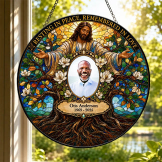 Personalized stained glass memorial hanging sign featuring Jesus Christ with outstretched arms, white flowers, a tree of life motif, and a central portrait photo of a man named Otis Anderson with the text 'Resting in Peace, Remembered in Love' and 'Otis Anderson 1969 - 2025'. The sign is hanging in front of a window with outdoor greenery visible.