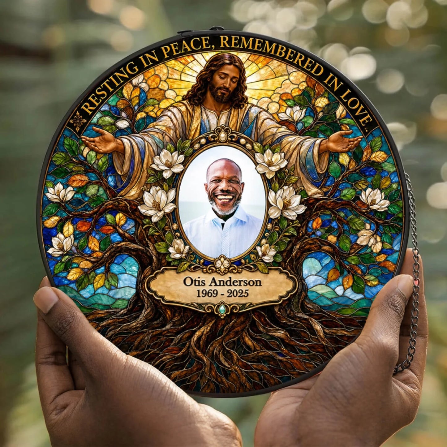 A circular memorial plaque or keepsake plate being held by two hands, featuring a stained-glass-style design with Jesus, magnolia flowers, tree roots, and a portrait of Otis Anderson with the text 'Resting in Peace, Remembered in Love' and 'Otis Anderson 1969 - 2025'.