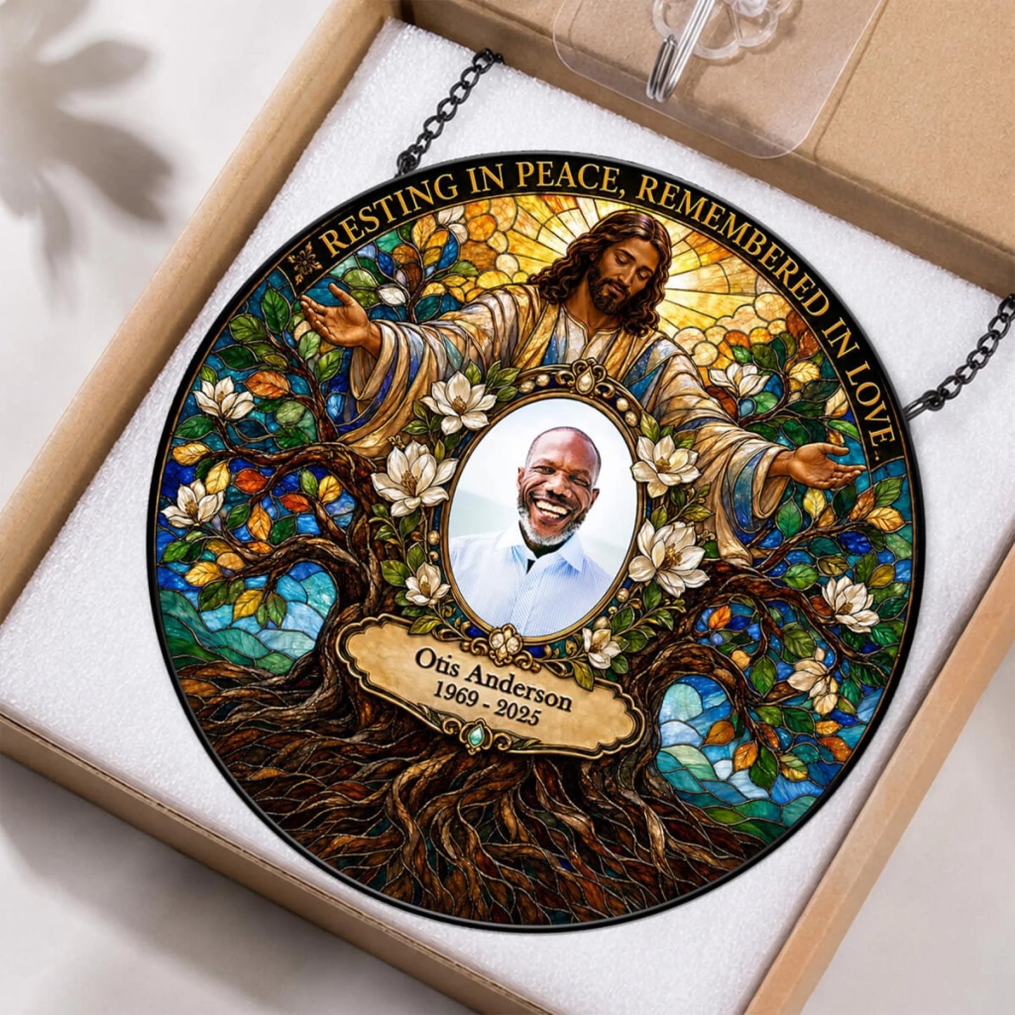 A personalized stained-glass-style memorial ornament displayed inside a gift box. The circular ornament features an illustrated figure of Jesus surrounded by white blossoms and a tree of life, with a central portrait photo and the text 'Resting in Peace, Remembered in Love' plus the name 'Otis Anderson 1969 - 2025'.