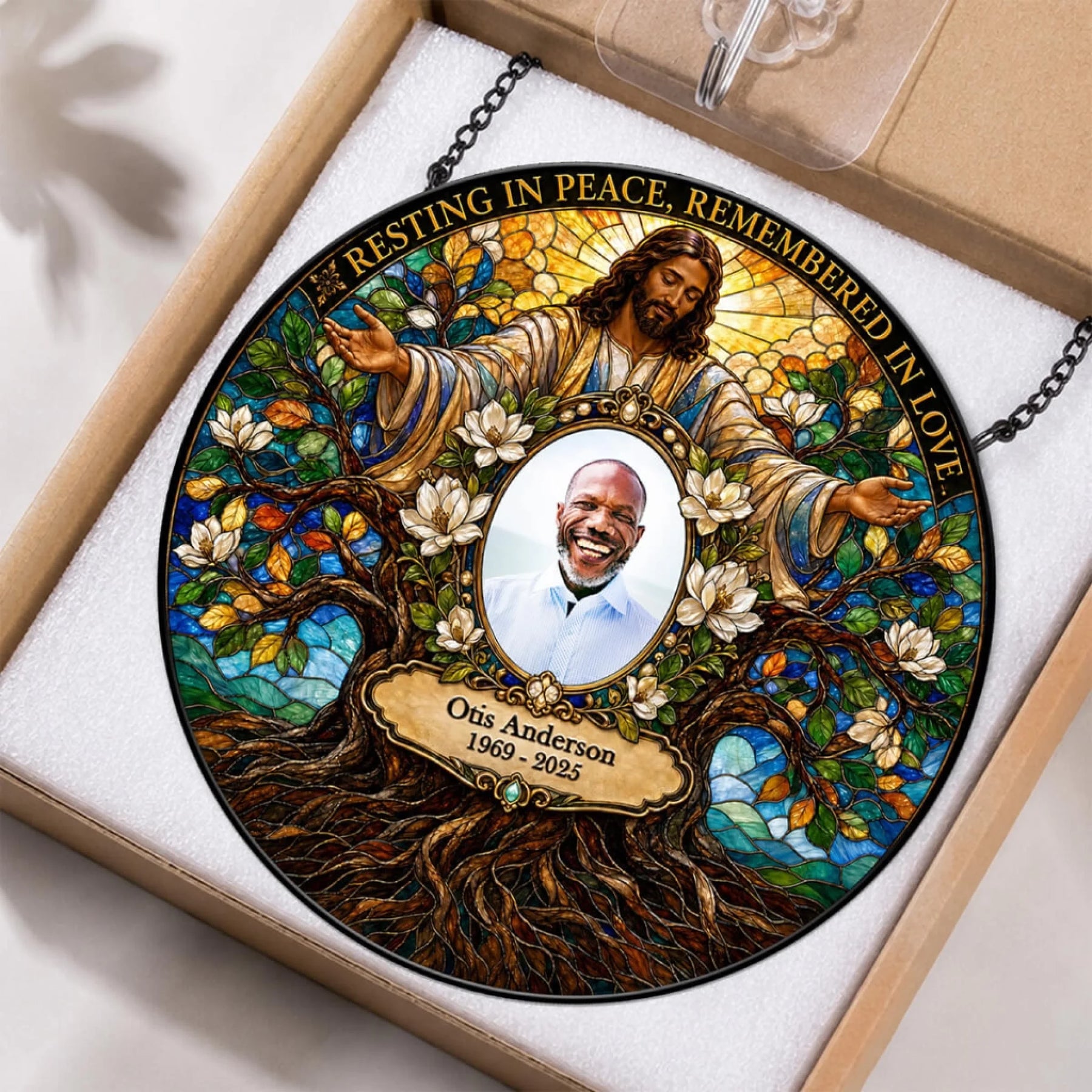 A personalized stained-glass-style memorial ornament displayed inside a gift box. The circular ornament features an illustrated figure of Jesus surrounded by white blossoms and a tree of life, with a central portrait photo and the text 'Resting in Peace, Remembered in Love' plus the name 'Otis Anderson 1969 - 2025'.