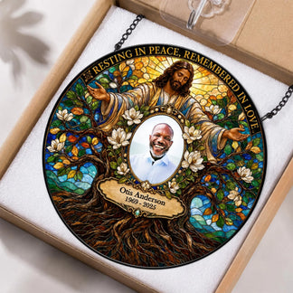 A personalized stained-glass-style memorial ornament displayed inside a gift box. The circular ornament features an illustrated figure of Jesus surrounded by white blossoms and a tree of life, with a central portrait photo and the text 'Resting in Peace, Remembered in Love' plus the name 'Otis Anderson 1969 - 2025'.