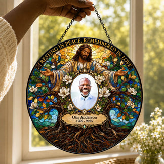 A personalized stained-glass-style hanging memorial plaque is held by a person's hand in front of a bright window. The circular design features Jesus at the top, white flowers, tree roots, and a central portrait of Otis Anderson with the text 'Resting in Peace, Remembered in Love' and 'Otis Anderson 1969 - 2025'.