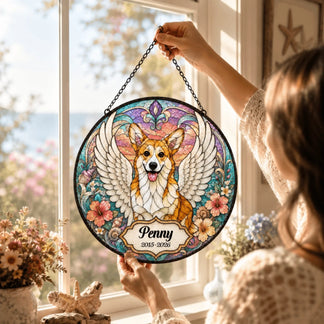 A person holds a circular stained glass style art piece featuring a winged corgi with a personalized name and dates, in a window.