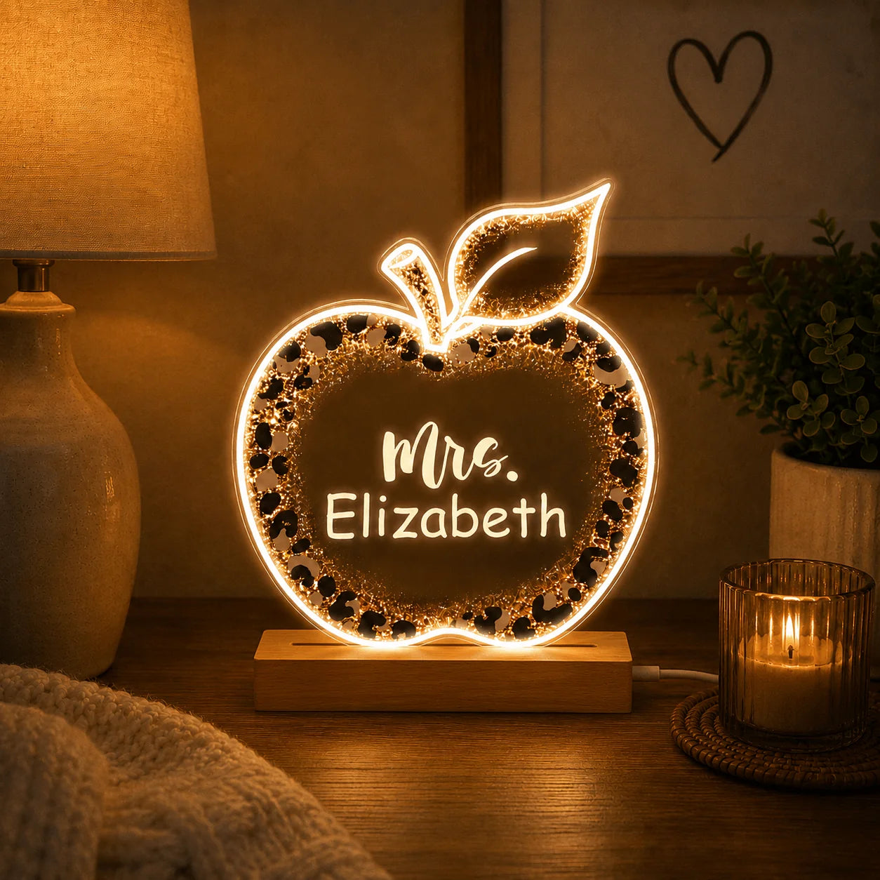 Decorative apple-shaped night light with 'Mrs. Elizabeth' on a wooden base, surrounded by a cozy setting with a lamp and candle.
