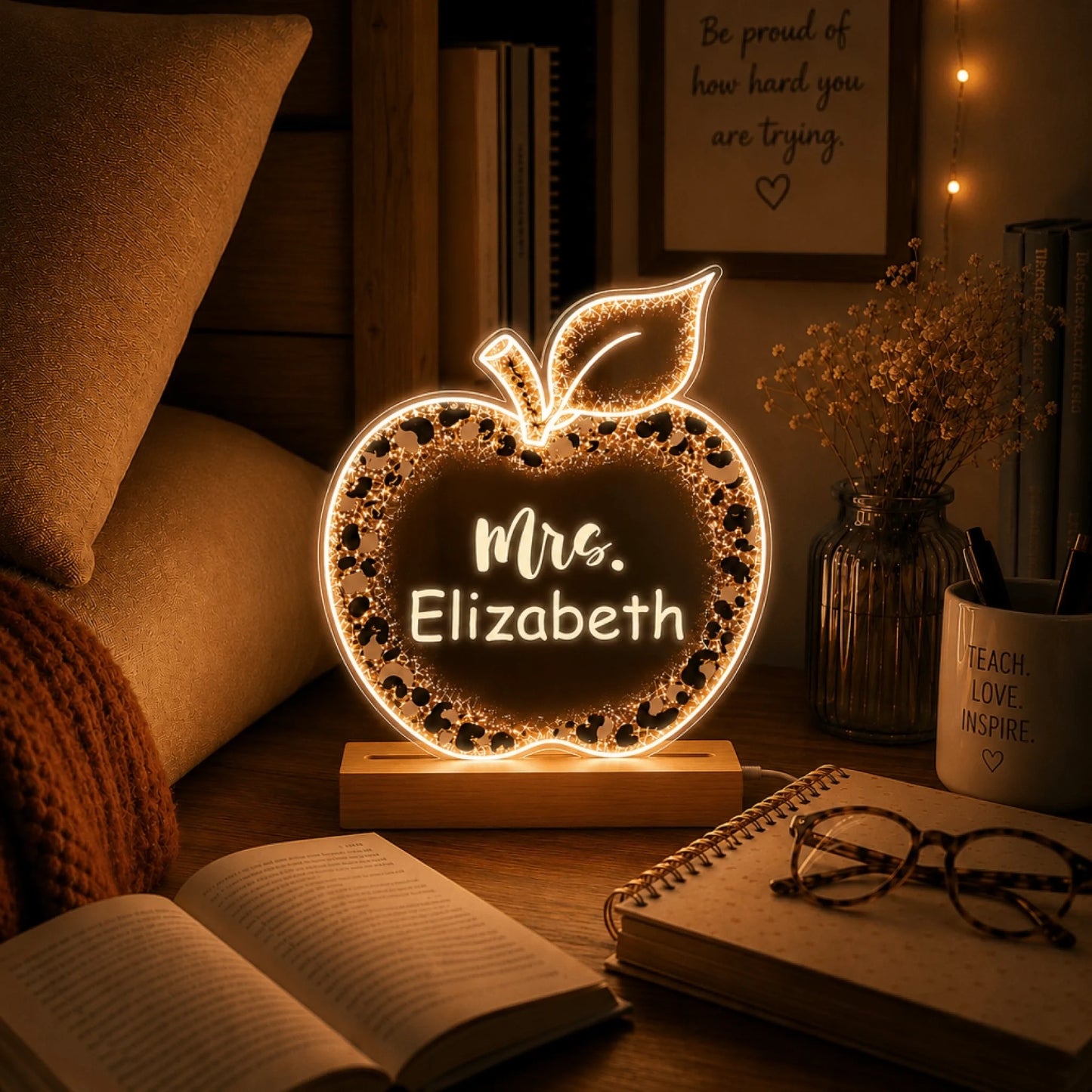 A glowing apple-shaped night light with a leopard print border and "Mrs. Elizabeth" text, on a desk with an open book and glasses.