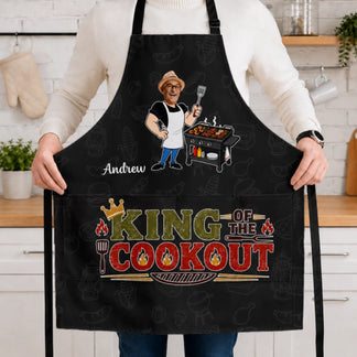 Person wearing a black apron with 'King of the Cookout' design in a kitchen.