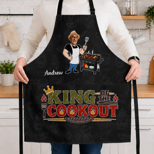 Person wearing a black apron with 'King of the Cookout' design in a kitchen.
