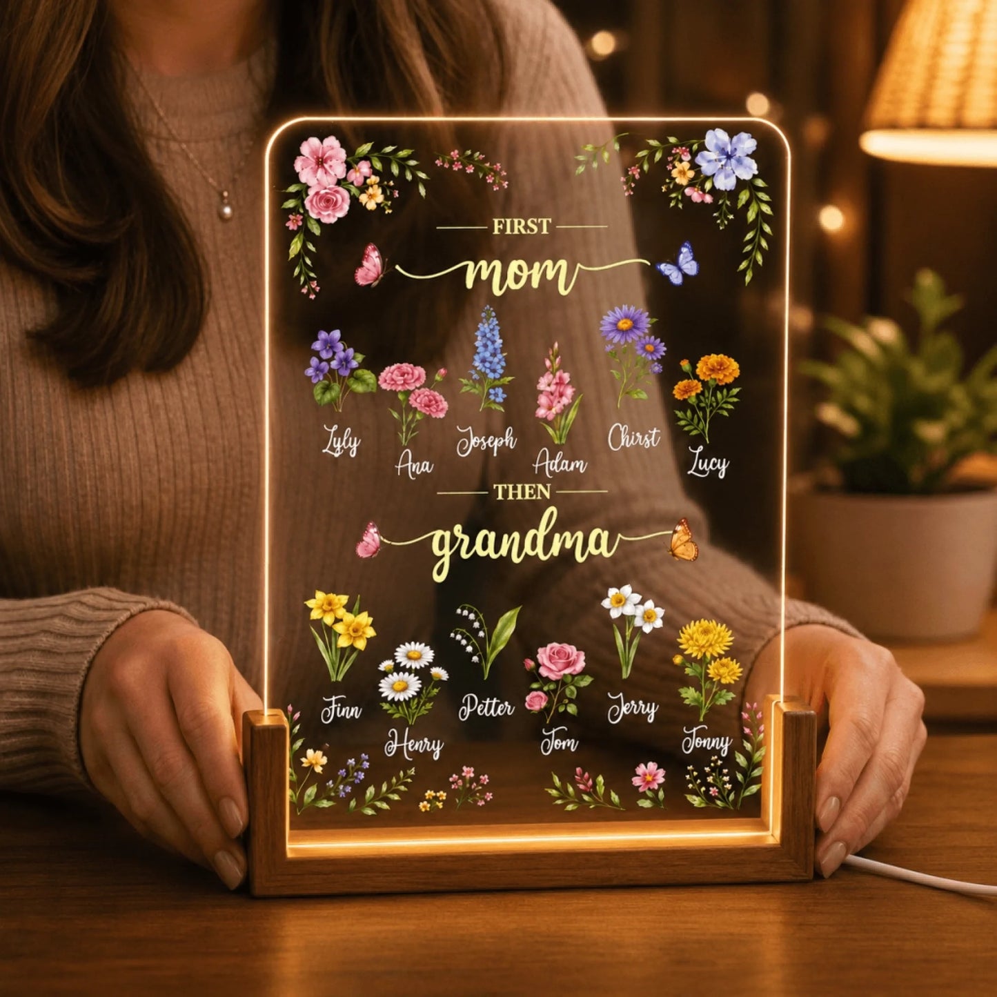 A person holds a glowing acrylic plaque that reads "FIRST mom THEN grandma," decorated with colorful flowers, names, and butterflies.