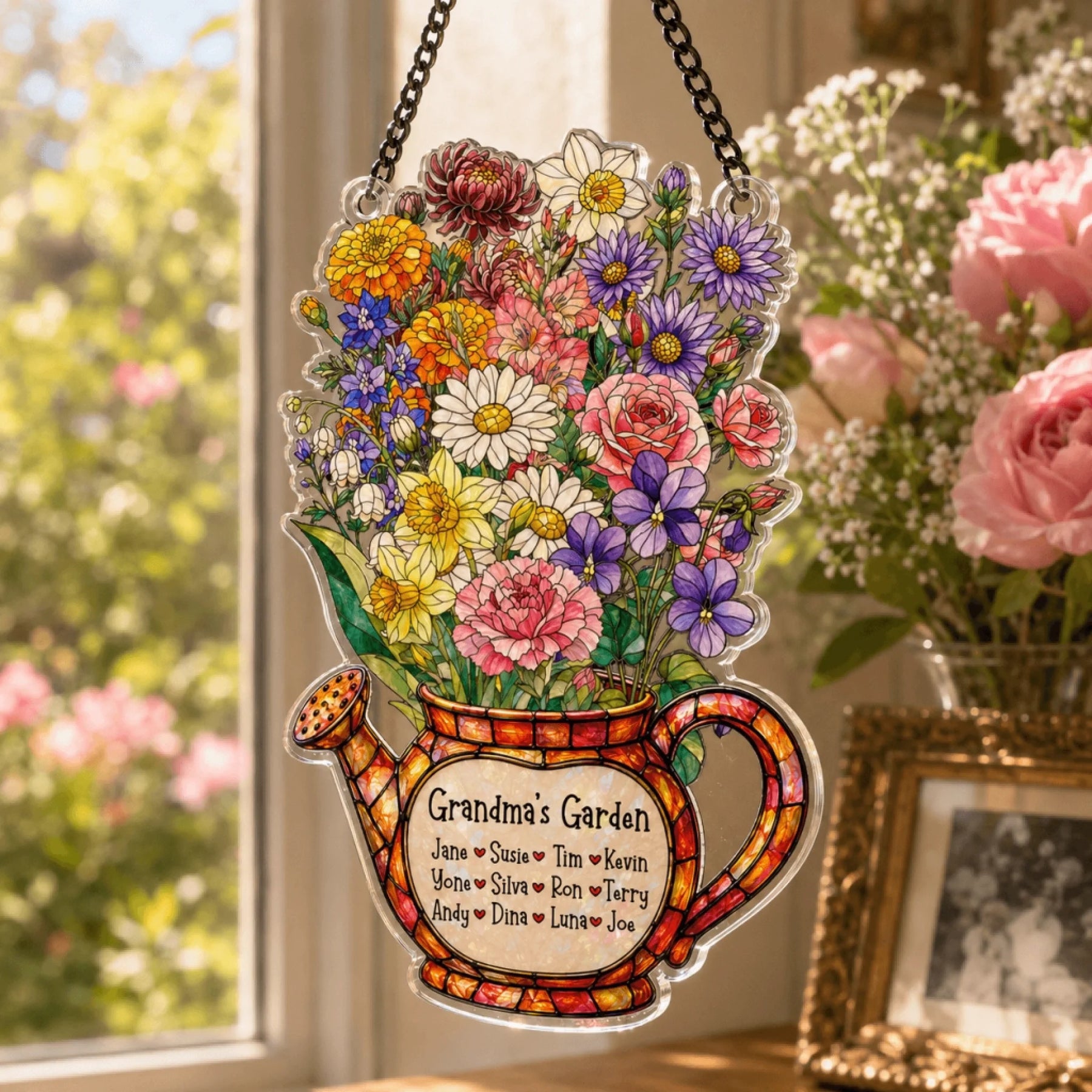 A colorful watering can decoration, overflowing with flowers and a 'Grandma's Garden' label, hangs in a sunlit window beside a lush floral bouquet.