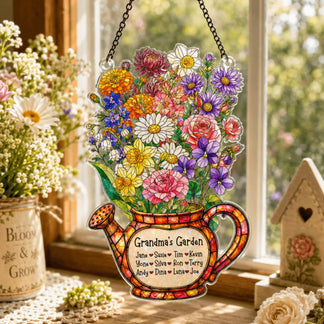 A colorful watering can shaped decoration, adorned with a vibrant bouquet of flowers and a 'Grandma's Garden' label, hangs by a chain in a sunlit window.