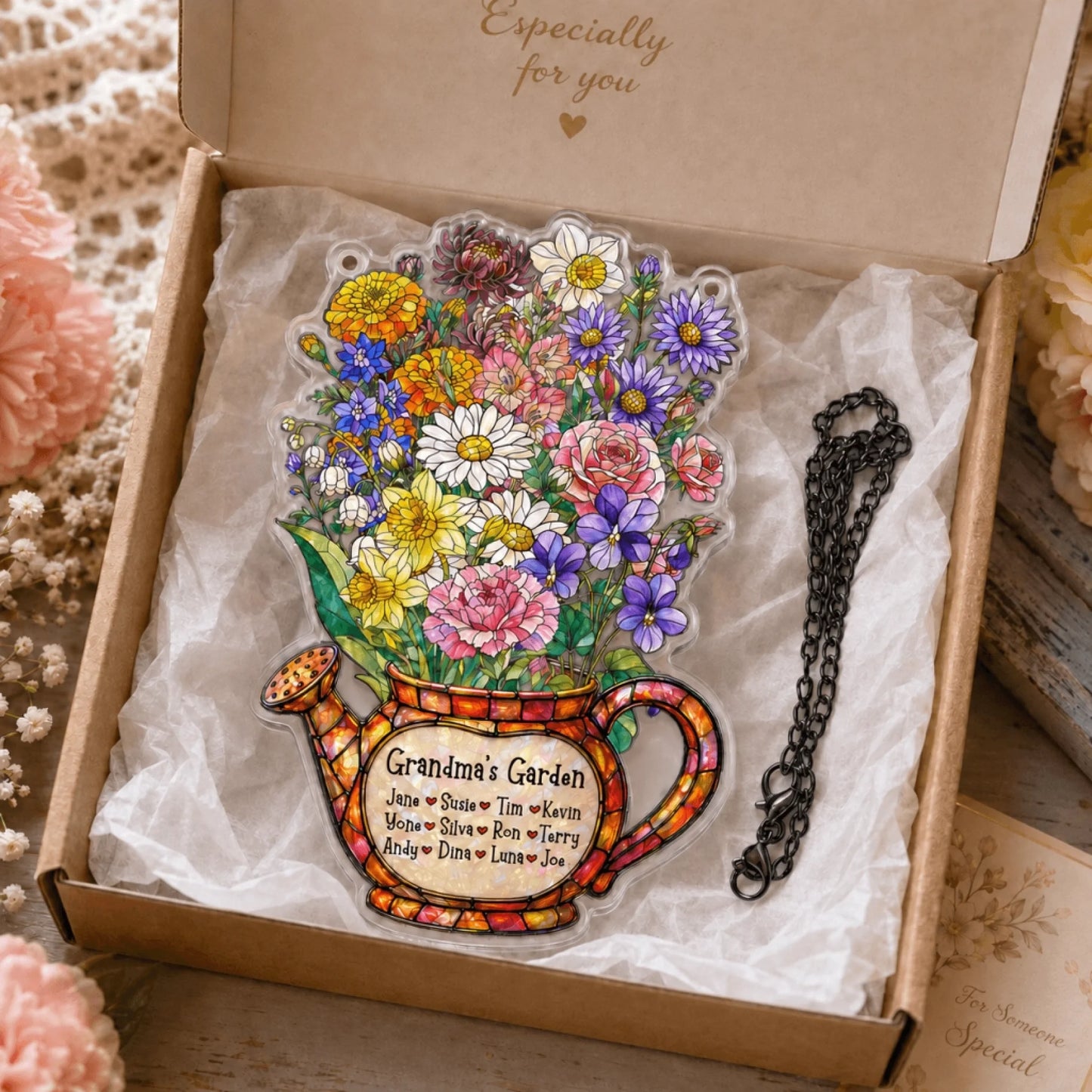 A clear acrylic watering can decoration filled with a colorful floral bouquet, detailed with a "Grandma's Garden" label and names. It sits in a gift box with a black chain beside it.