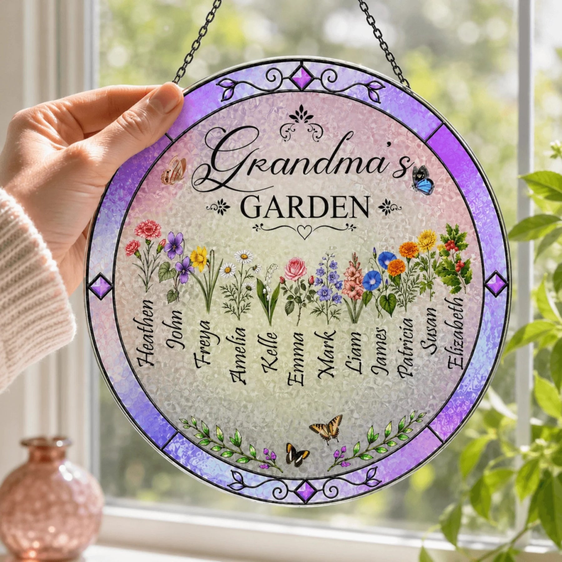 Round stained glass panel with floral design and names, held by a hand in front of a window with plants.