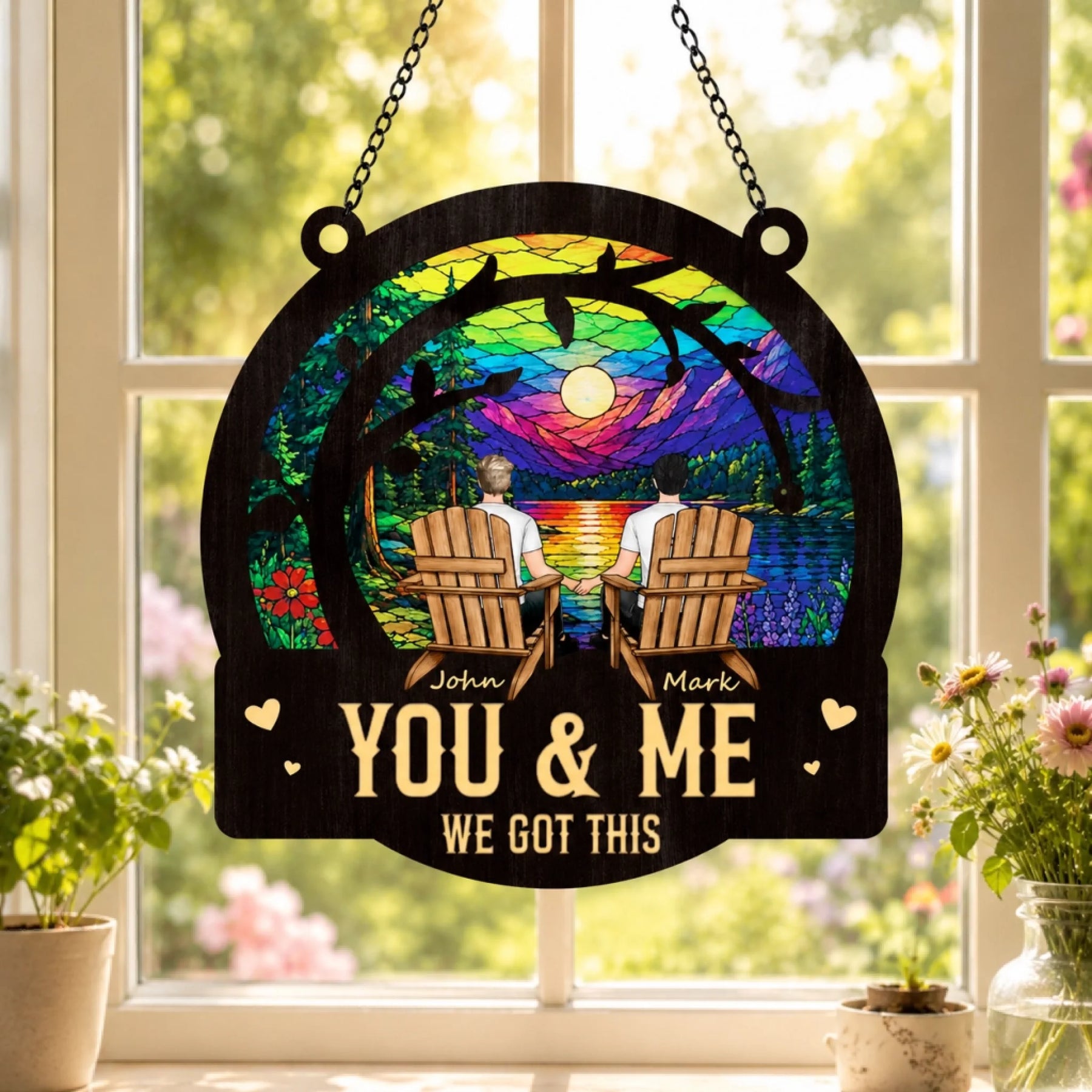 Decorative sign with 'You & Me We Got This' text, two people in chairs, and a rainbow sunset design, hanging in front of a window.