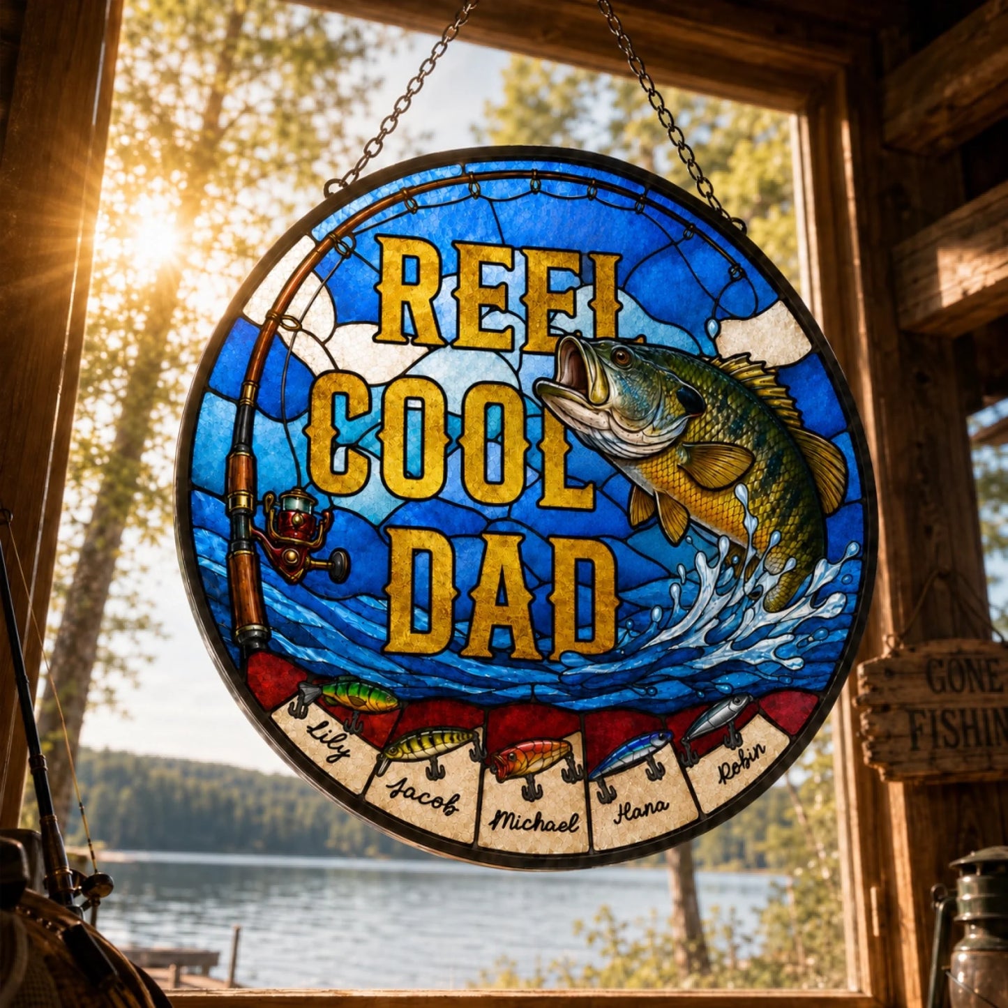 Stained glass panel with 'Reef Cool Dad' text and fish design, hanging by a window with a view of trees and water.