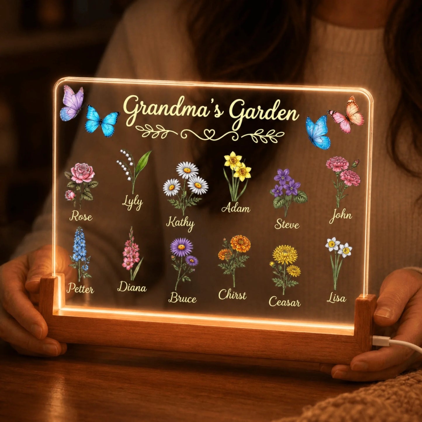 Illuminated acrylic plaque features 'Grandma's Garden' text, colorful butterflies, and a grid of various flowers, each paired with a name. It stands on a wooden light-up base.