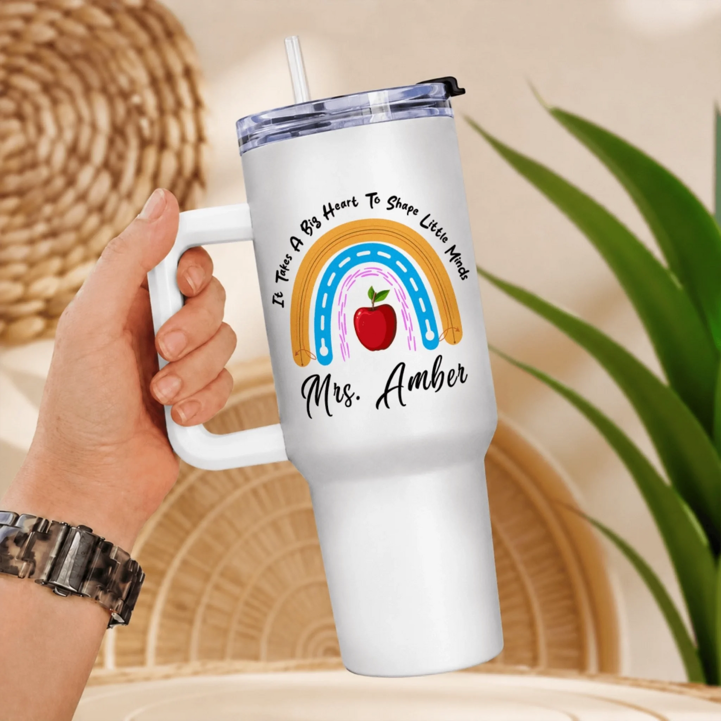 A hand holding a white handled tumbler with a rainbow, red apple, and text design, against a blurred background of woven patterns.