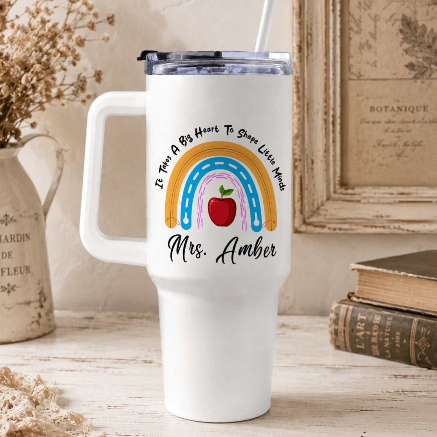 A white handled tumbler with a rainbow and apple design on a rustic desk with dried flowers, old books, and a framed botanical print.
