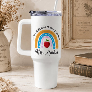 A white handled tumbler with a rainbow and apple design on a rustic desk with dried flowers, old books, and a framed botanical print.
