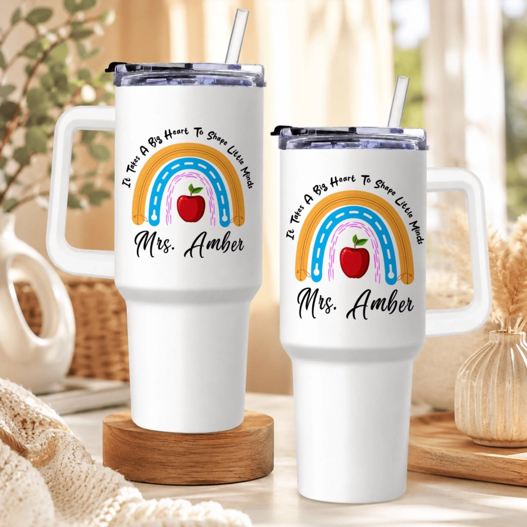 Two white handled tumblers, both with the same rainbow, red apple, and text design, sitting on a table with a knitted throw.